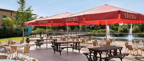 Un patio al aire libre con mesas y sillas, sombrillas rojas y una fuente de fondo.