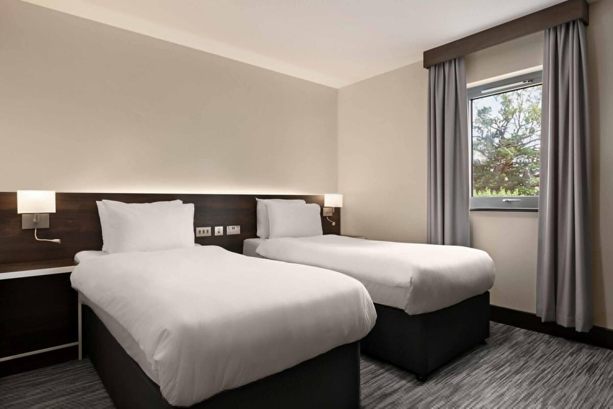 A hotel room with two beds, a nightstand with a lamp, a window with curtains, and a view of trees outside.