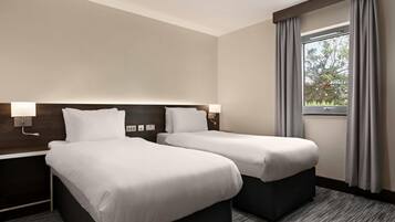 Habitación de hotel con dos camas, una mesita de noche con lámpara, ventana con cortinas y vista a árboles.