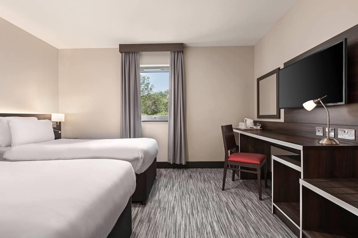 A hotel room with two beds, a desk, a chair, a TV, and a window with curtains.