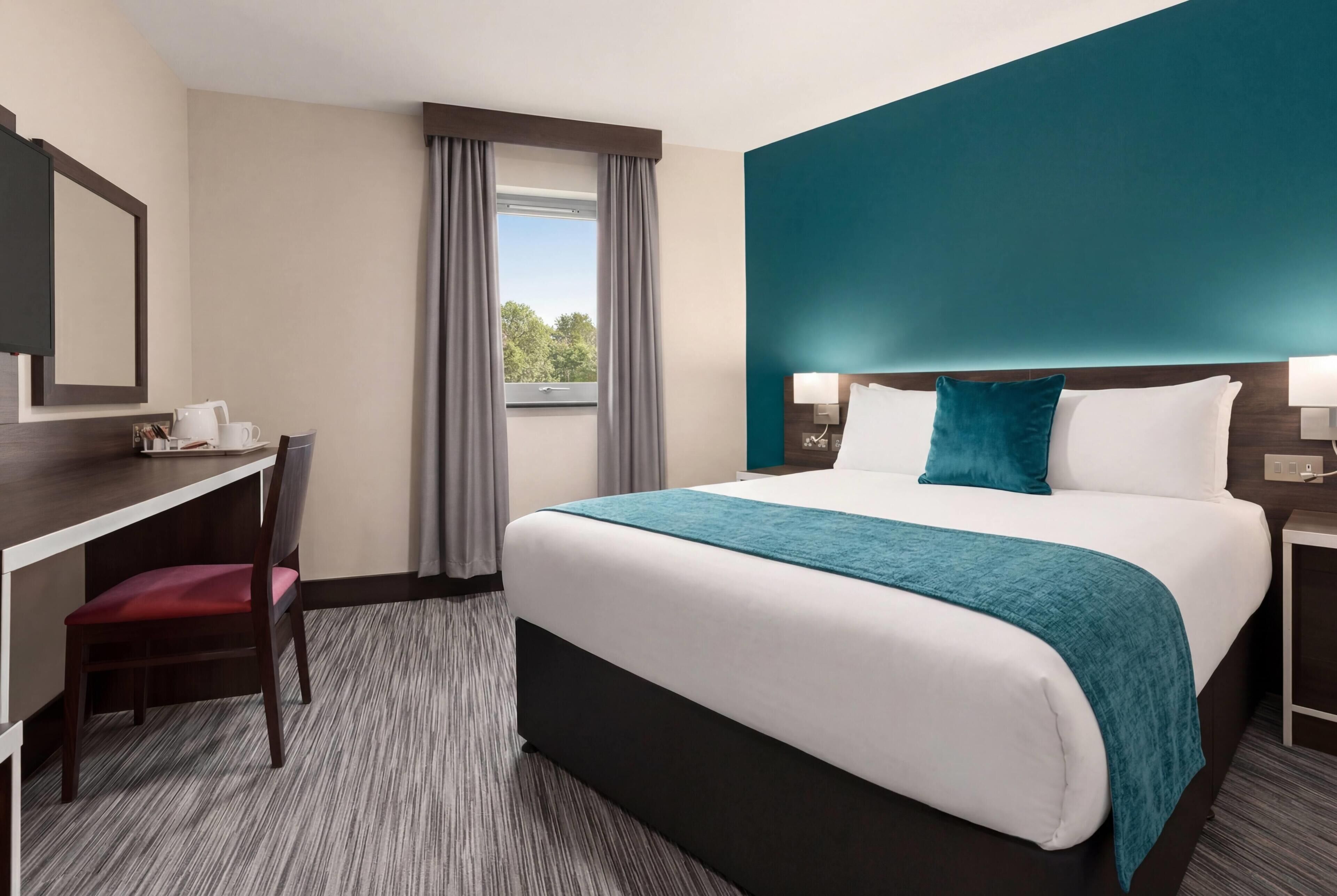 A hotel room with a bed, a desk, a chair, a window with curtains, and a view of trees.
