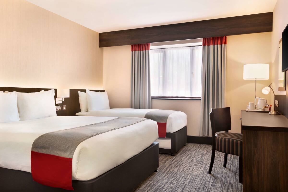 A hotel room with two beds, a desk, a chair, and a window with curtains.