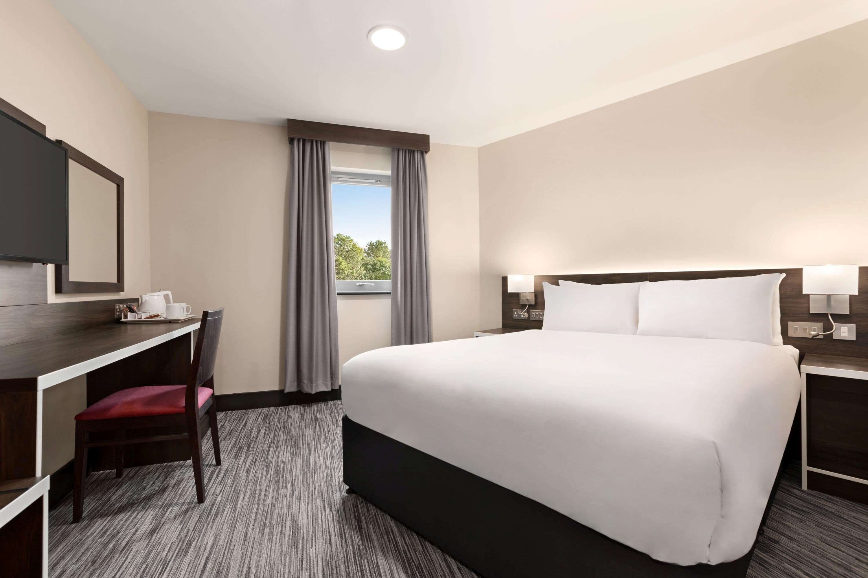 Une chambre d’hôtel comprenant un lit, un bureau avec une chaise, une fenêtre avec des rideaux et une table de chevet avec une lampe.