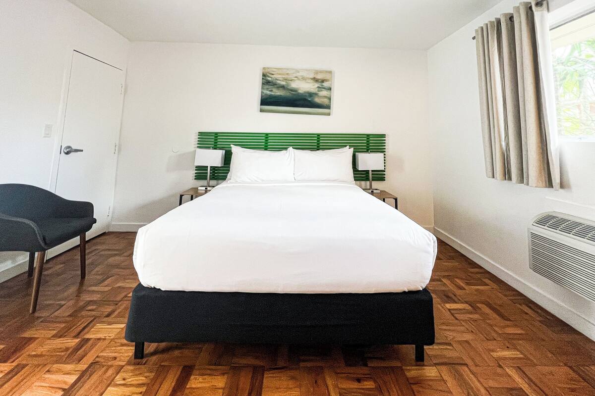 Une chambre d’hôtel avec un grand lit, une tête de lit verte, deux tables de chevet avec des lampes, une chaise, un tableau au mur et une fenêtre avec des rideaux.