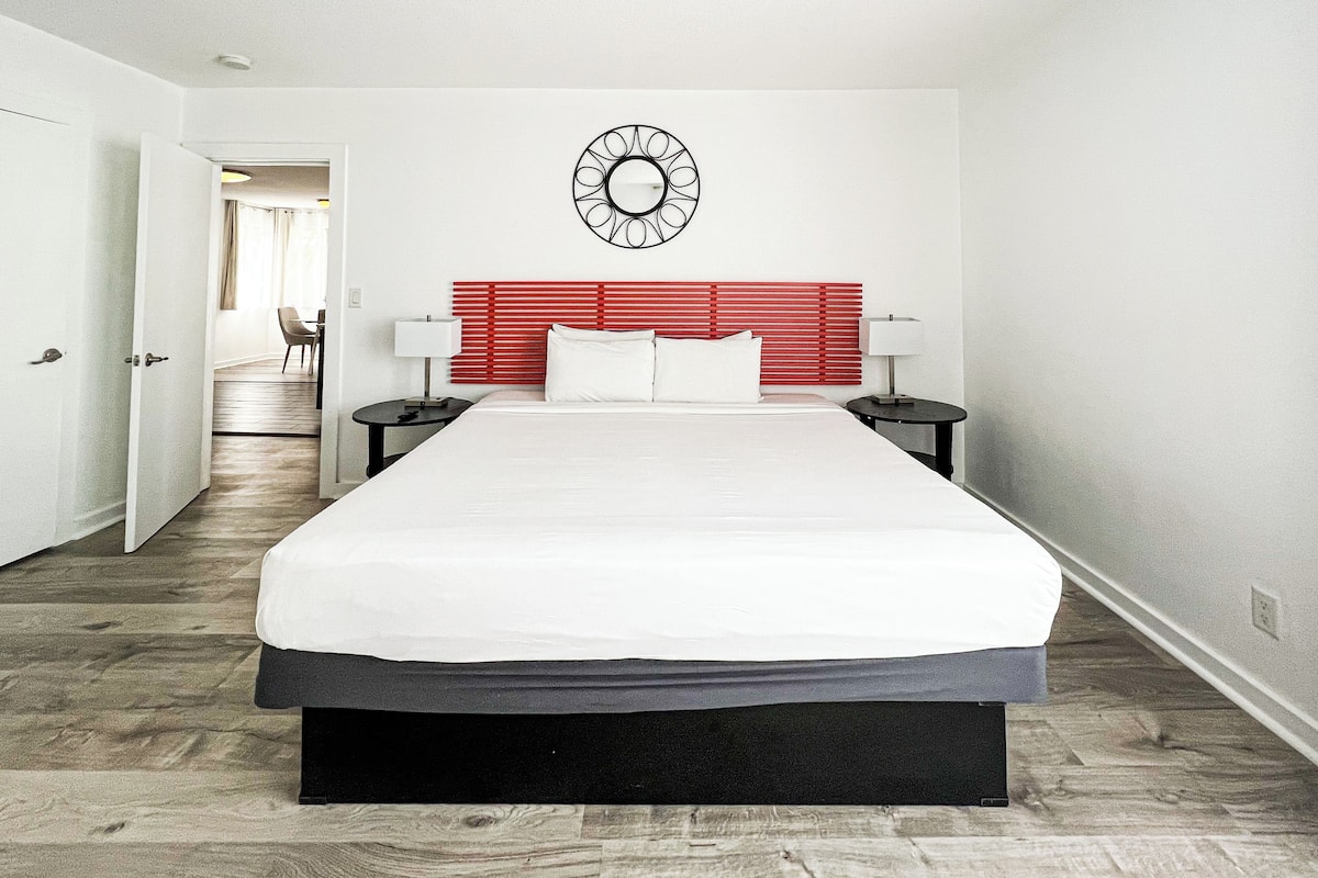 Une chambre d’hôtel moderne avec un grand lit, une tête de lit rouge, deux tables de chevet avec des lampes et une horloge murale.