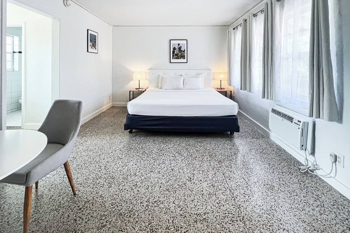 Une chambre d’hôtel comprenant un lit, deux tables de chevet avec des lampes, une chaise, une table, un ventilateur de plafond et une fenêtre avec des rideaux.