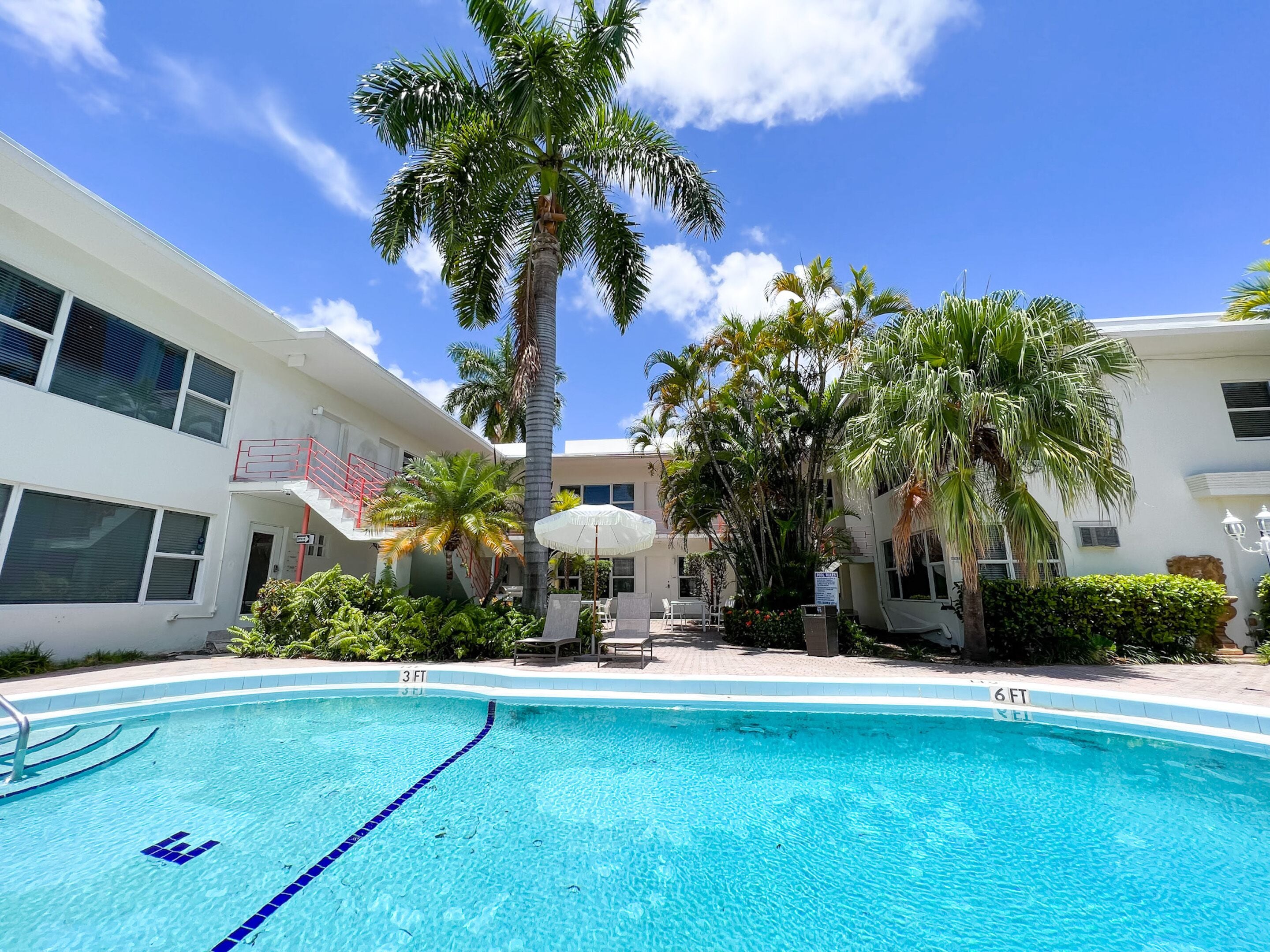 A swimming pool with clear blue water, surrounded by palm trees and a white building with multiple windows and balconies.