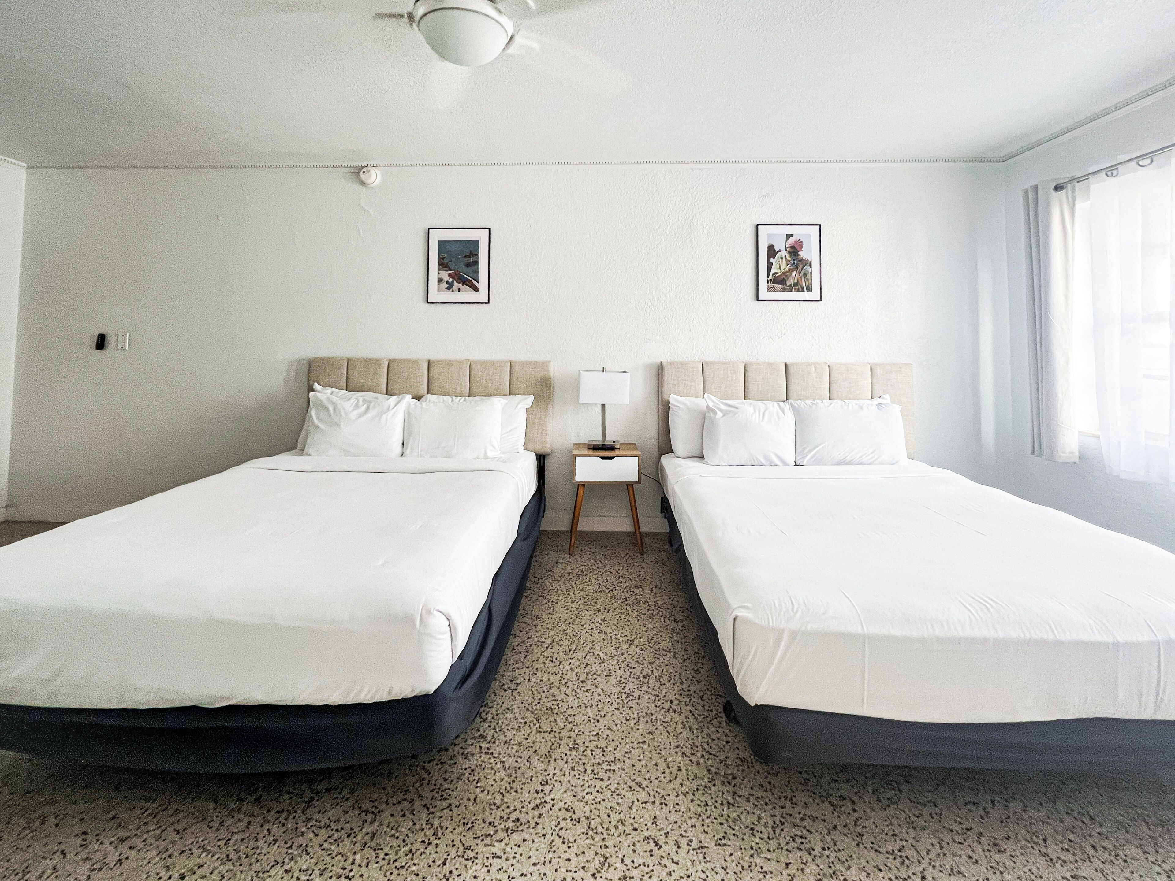 Ein Hotelzimmer mit zwei Betten, einem Deckenventilator, einem kleinen Nachttisch mit Lampe und zwei gerahmten Bildern an der Wand.