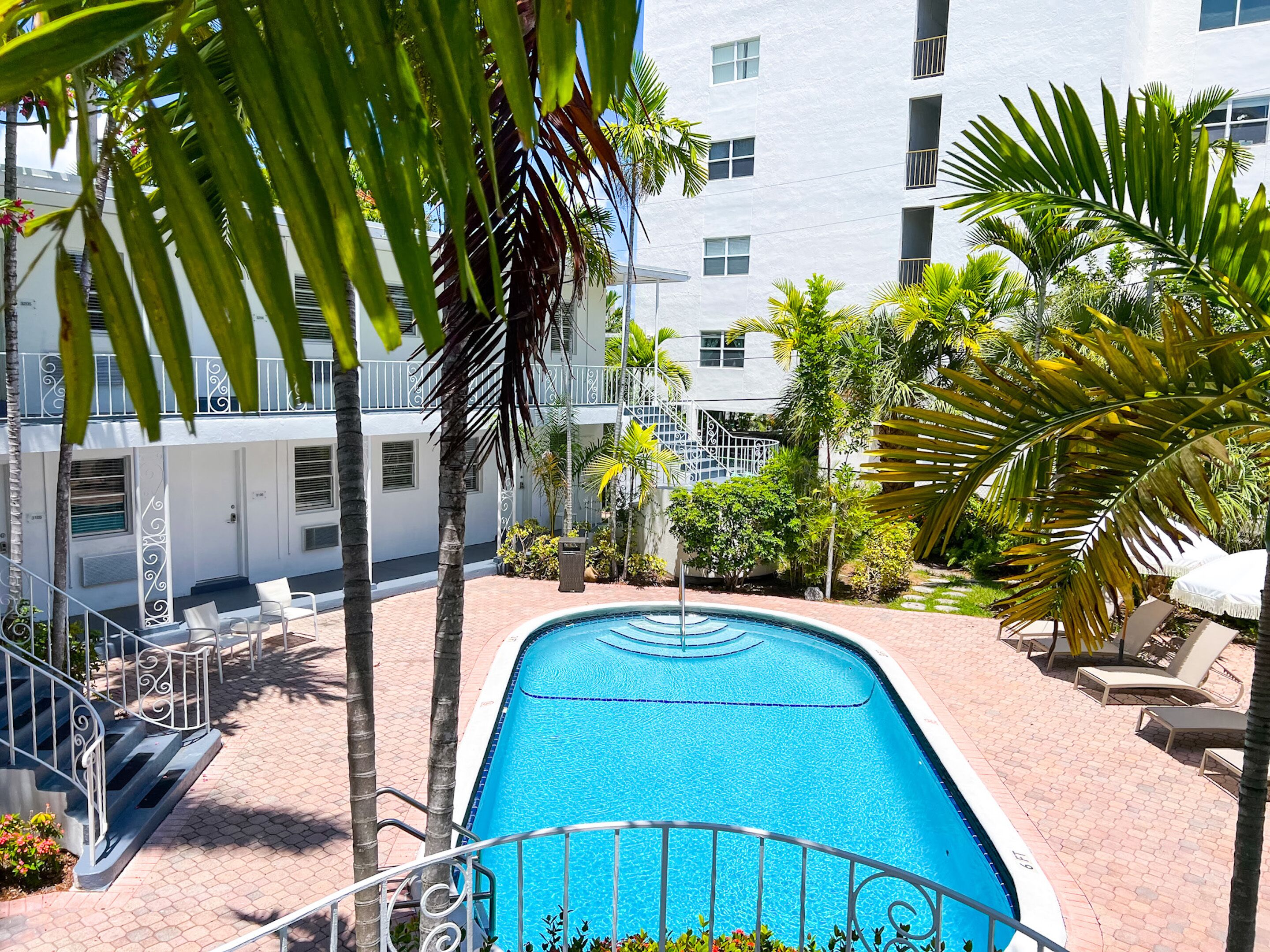 A swimming pool with a metal railing, surrounded by palm trees and a white building with balconies.