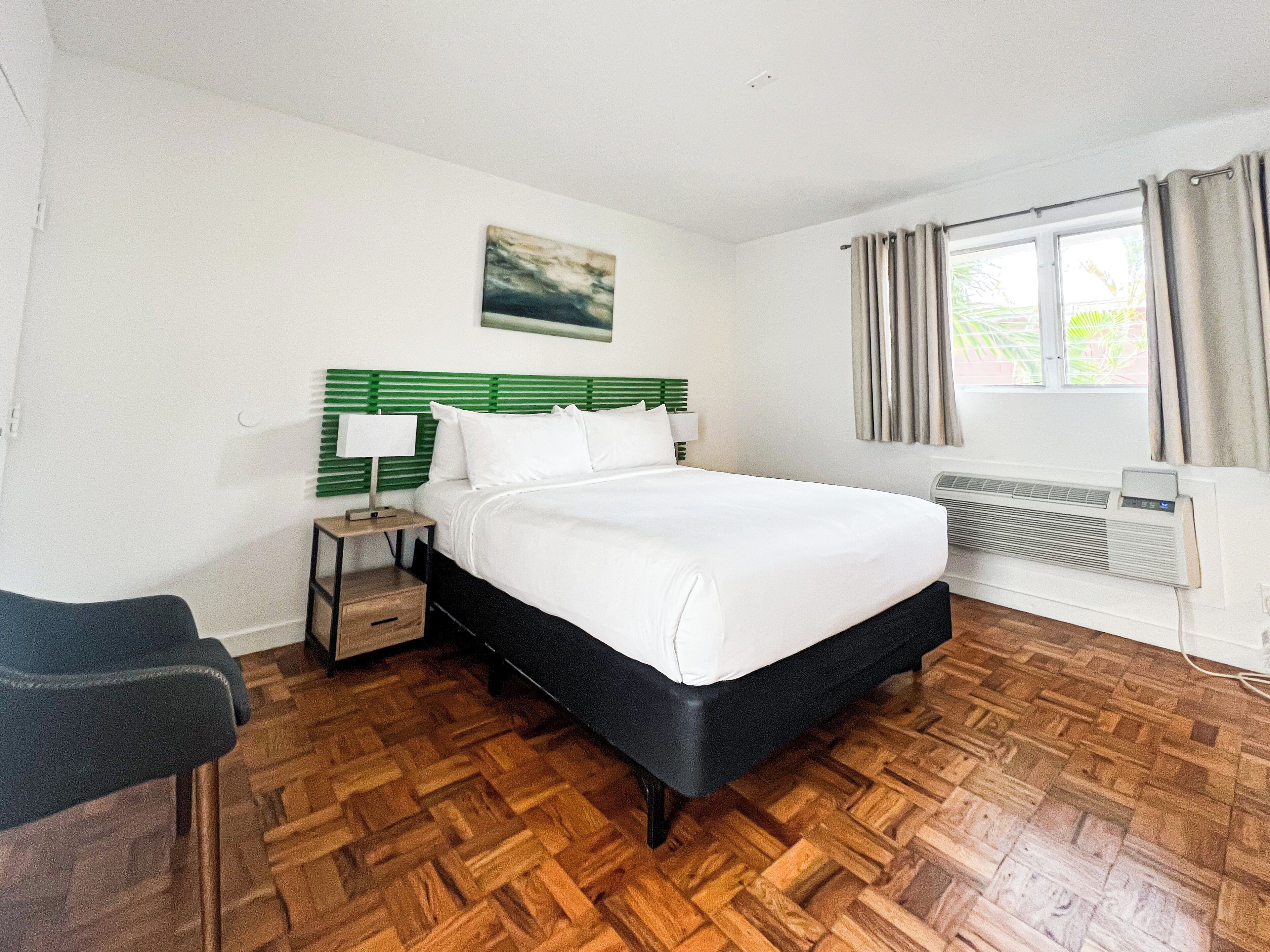 A hotel room with a bed, a nightstand, a chair, a window with curtains, and a radiator.