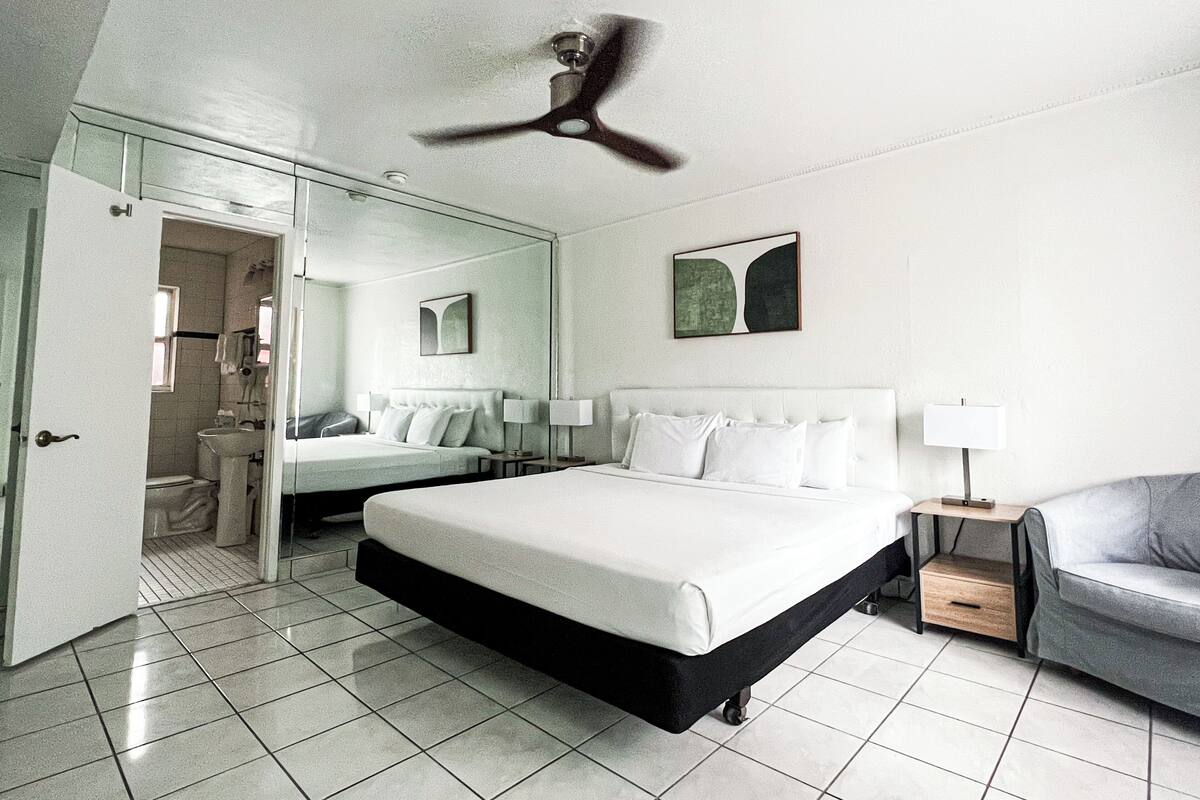 Une chambre d’hôtel moderne avec un grand lit, une table de chevet avec une lampe, une chaise, un ventilateur de plafond et une salle de bain visible à travers une porte ouverte.