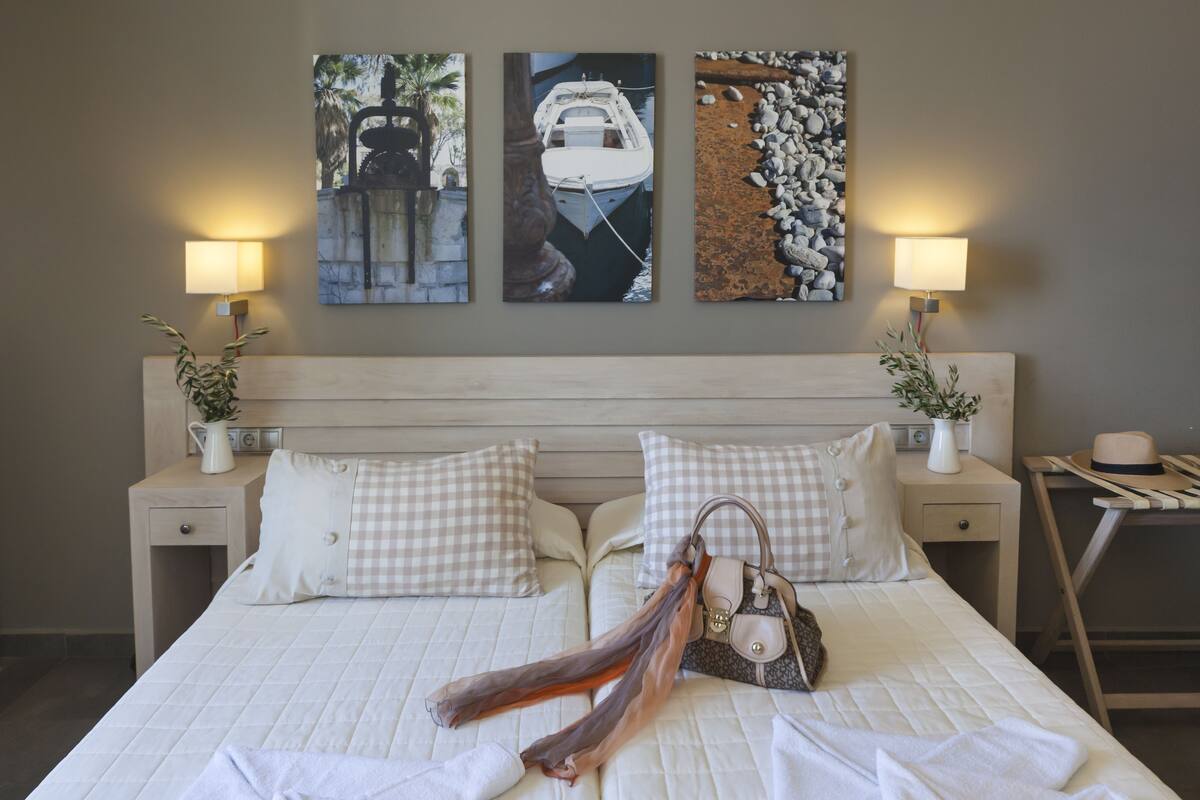 Cama bem arrumada com almofadas xadrez, um lenço decorativo e uma bolsa sobre a cama. Há duas mesas de cabeceira com abajures e uma mesinha com um chapéu e um vaso de flores.