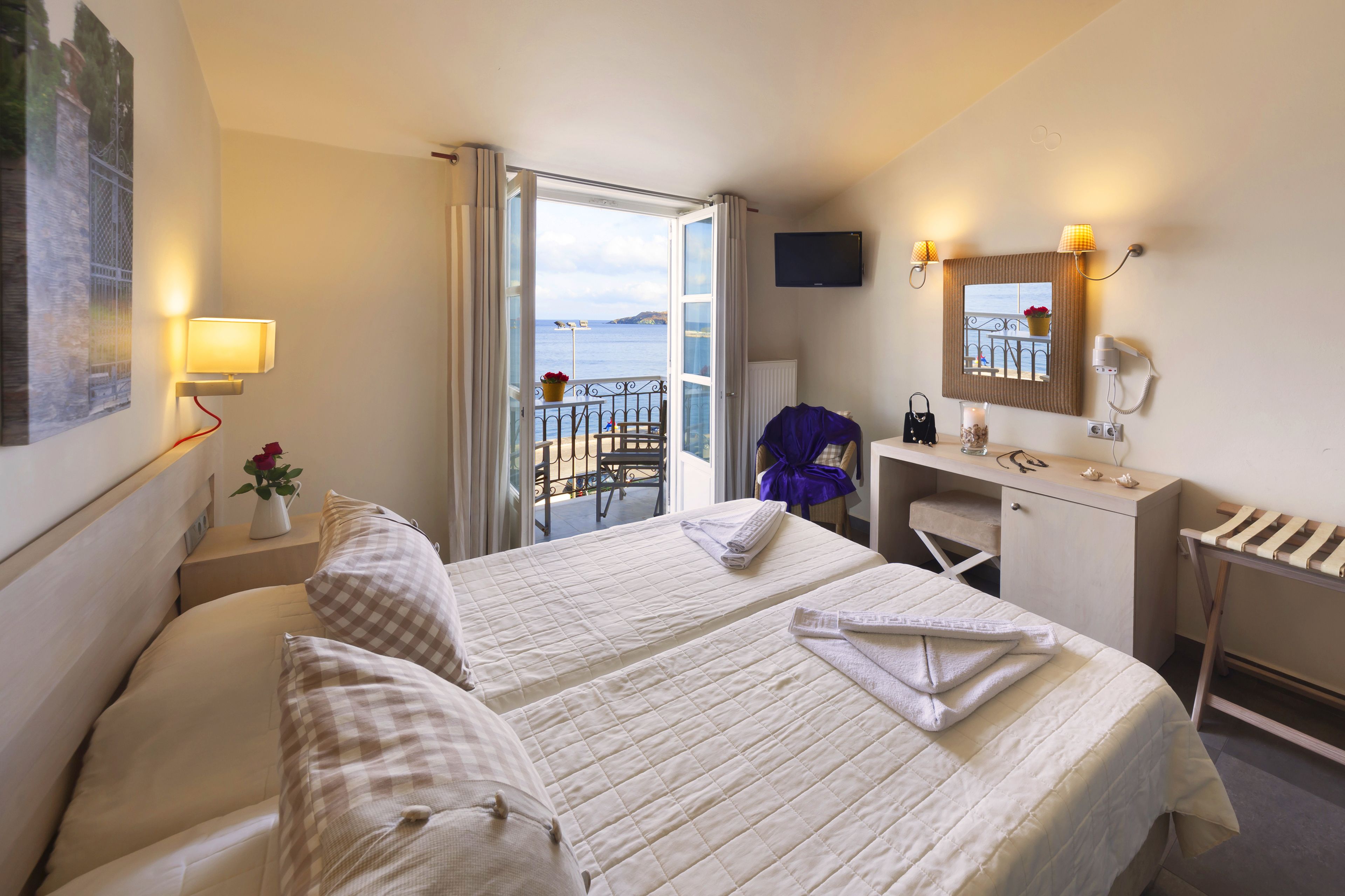 A hotel room with two beds, a desk, a chair, a small table with a vase, and a view of the sea through an open door.