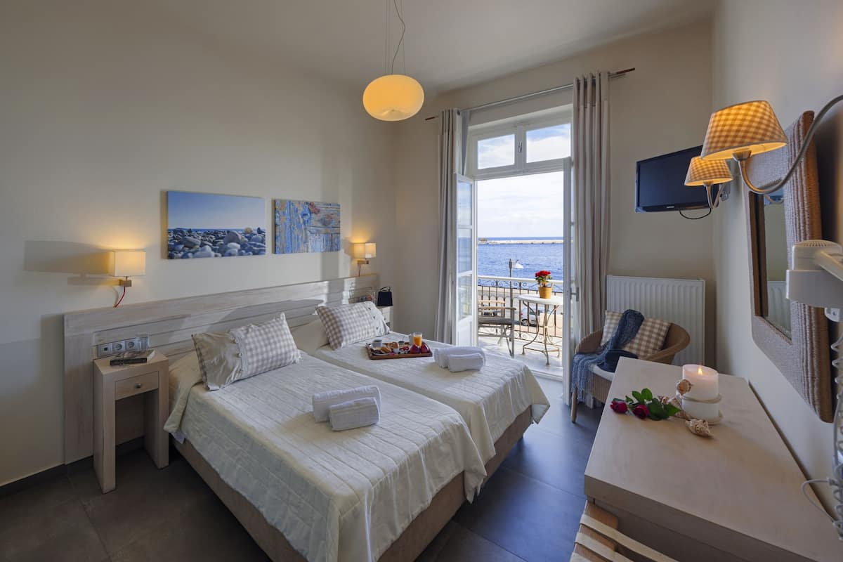 Quarto de hotel com duas camas, uma escrivaninha, uma cadeira, televisão e varanda com vista para o mar.