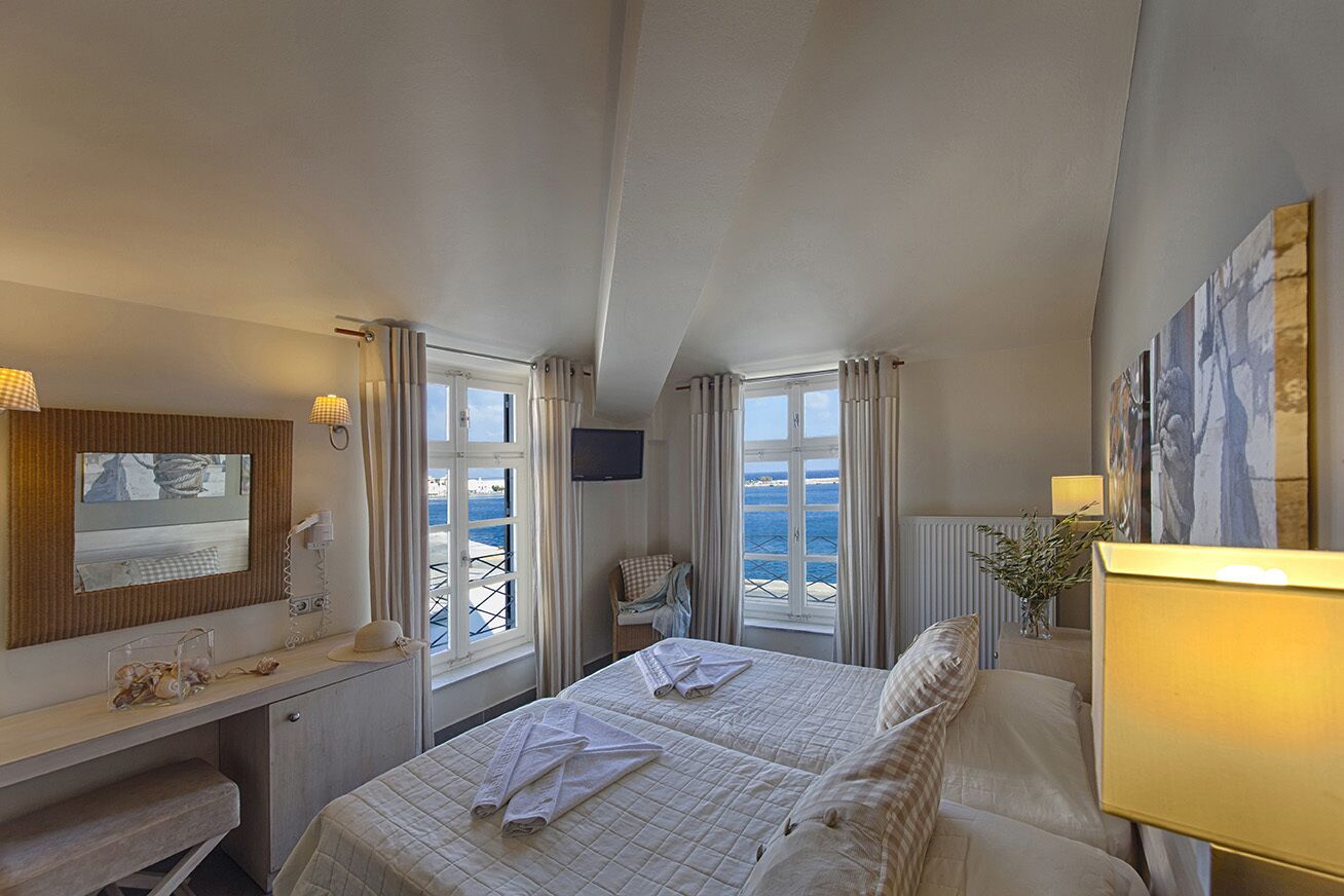 Ett hotellrum med två sängar, ett skrivbord, en stol, en TV och stora fönster med utsikt över havet.