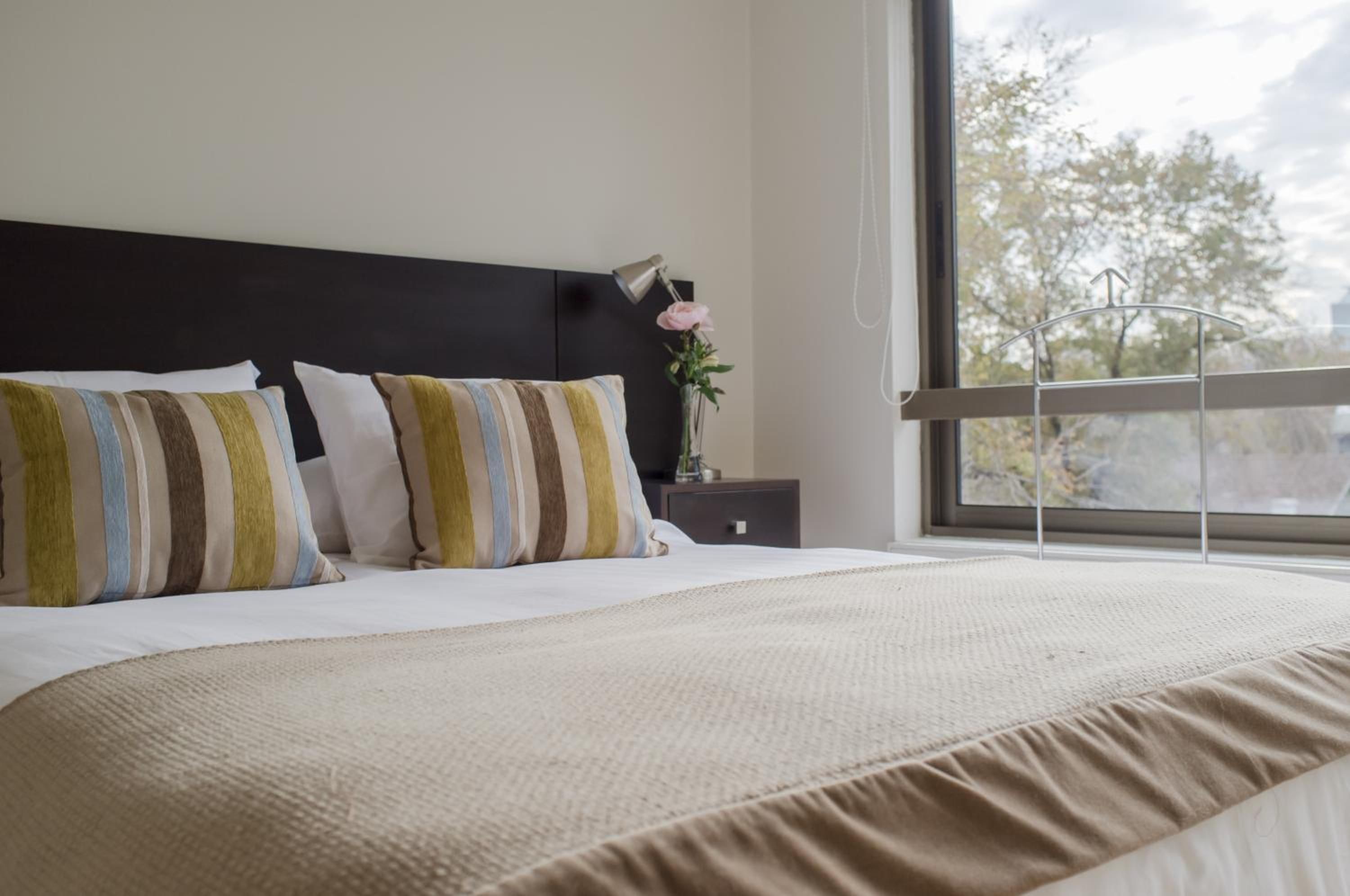 A neatly made bed with striped pillows, a bedside table with a lamp and a vase of flowers, and a window with a view of trees.