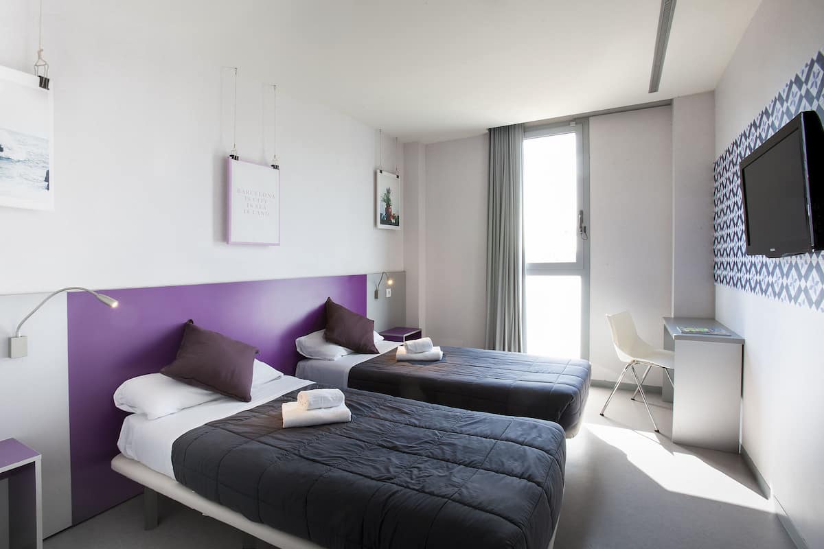 A modern hotel room with two beds, a desk, and a TV.