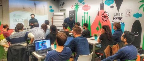 A classroom with students seated at desks, a presentation screen, and a colorful wall with various icons and text.