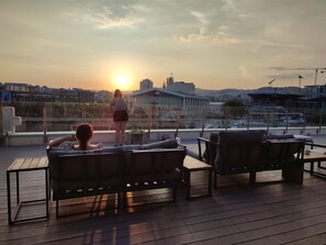 Une terrasse sur le toit avec deux chaises longues, une personne debout et une vue sur le coucher de soleil.