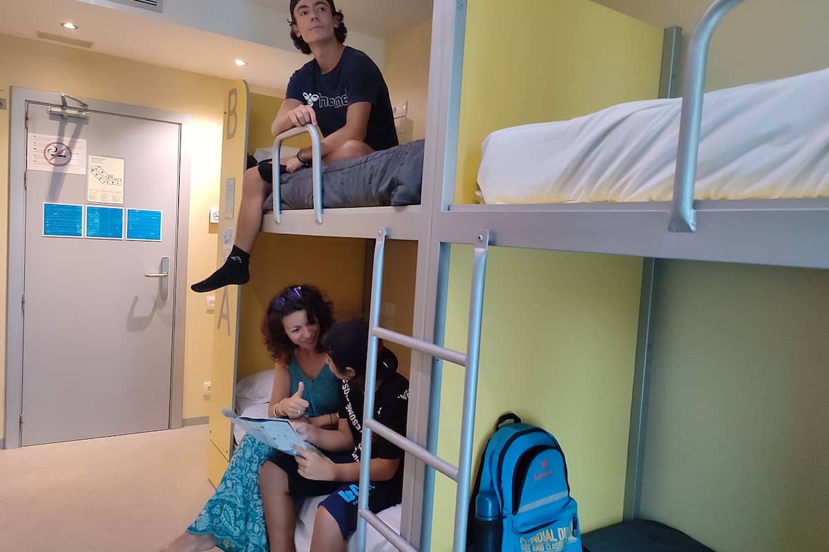 A bunk bed room with a door, a blue backpack, and a person reading a book.
