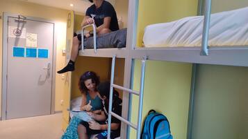 Une chambre avec un lit superposé, une porte, un sac à dos bleu et une personne qui lit un livre.