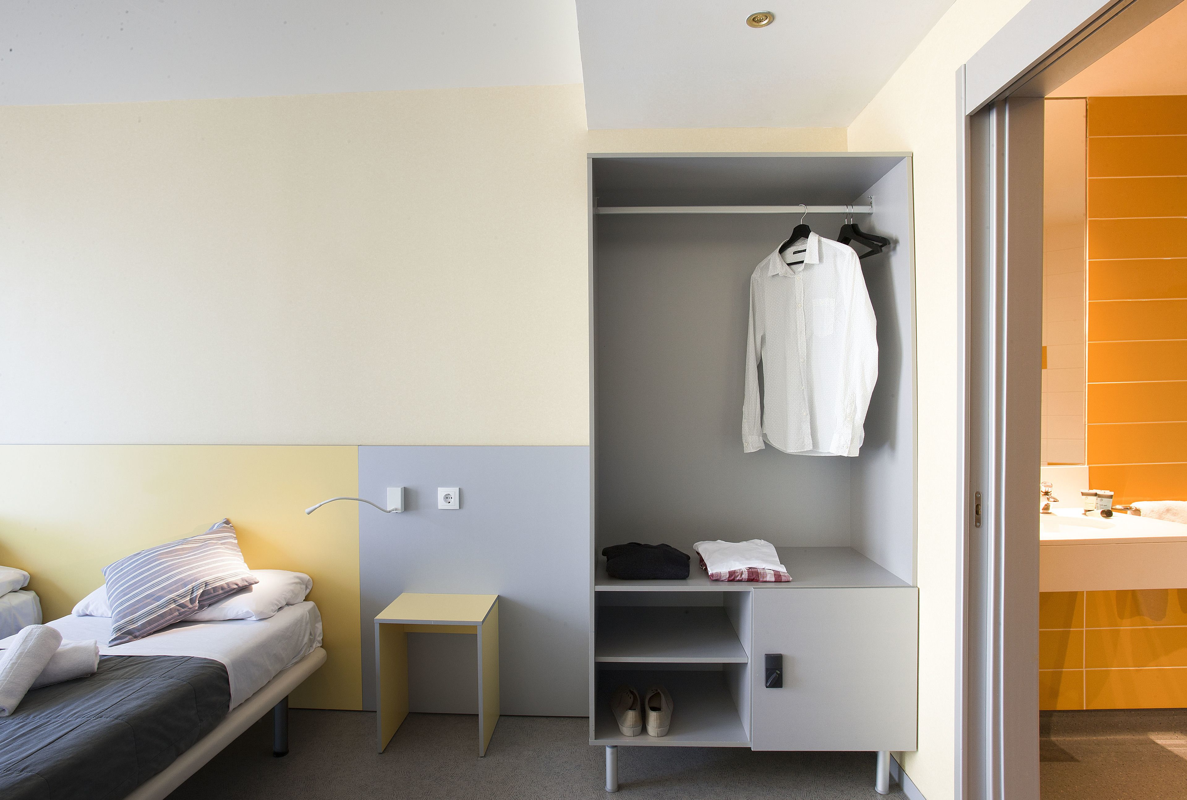 Ein modernes Hotelzimmer mit einem Bett, einem kleinen Tisch, einem Kleiderschrank und einem durch eine offene Tür einsehbarer Badezimmer.