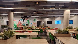 Una cafetería moderna con mesas de madera y sillas verdes, decorada con plantas y carteles motivacionales.