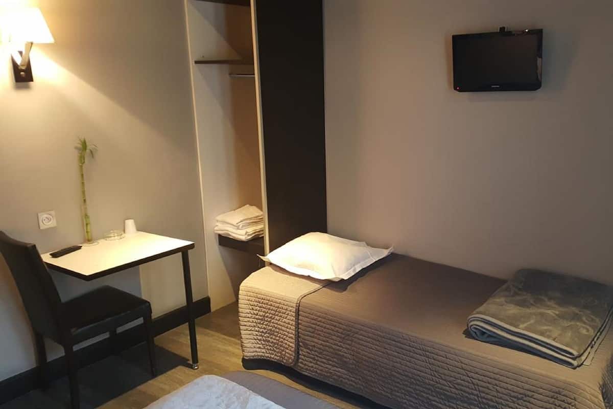 A small, neatly arranged bedroom with a single bed, a bedside table, a chair, a desk, and a TV mounted on the wall.
