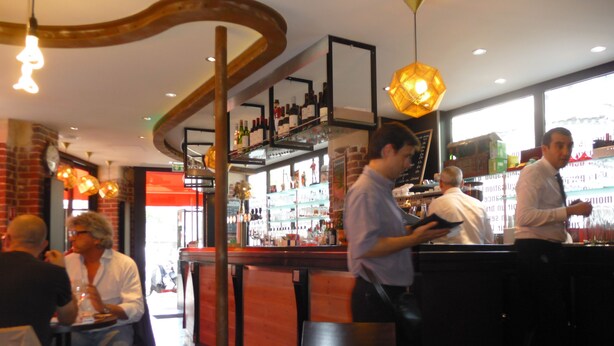 A bar area with a round table, shelves with bottles, and hanging lights. - Café hôtel de l'Avenir (Saint-Ouen)