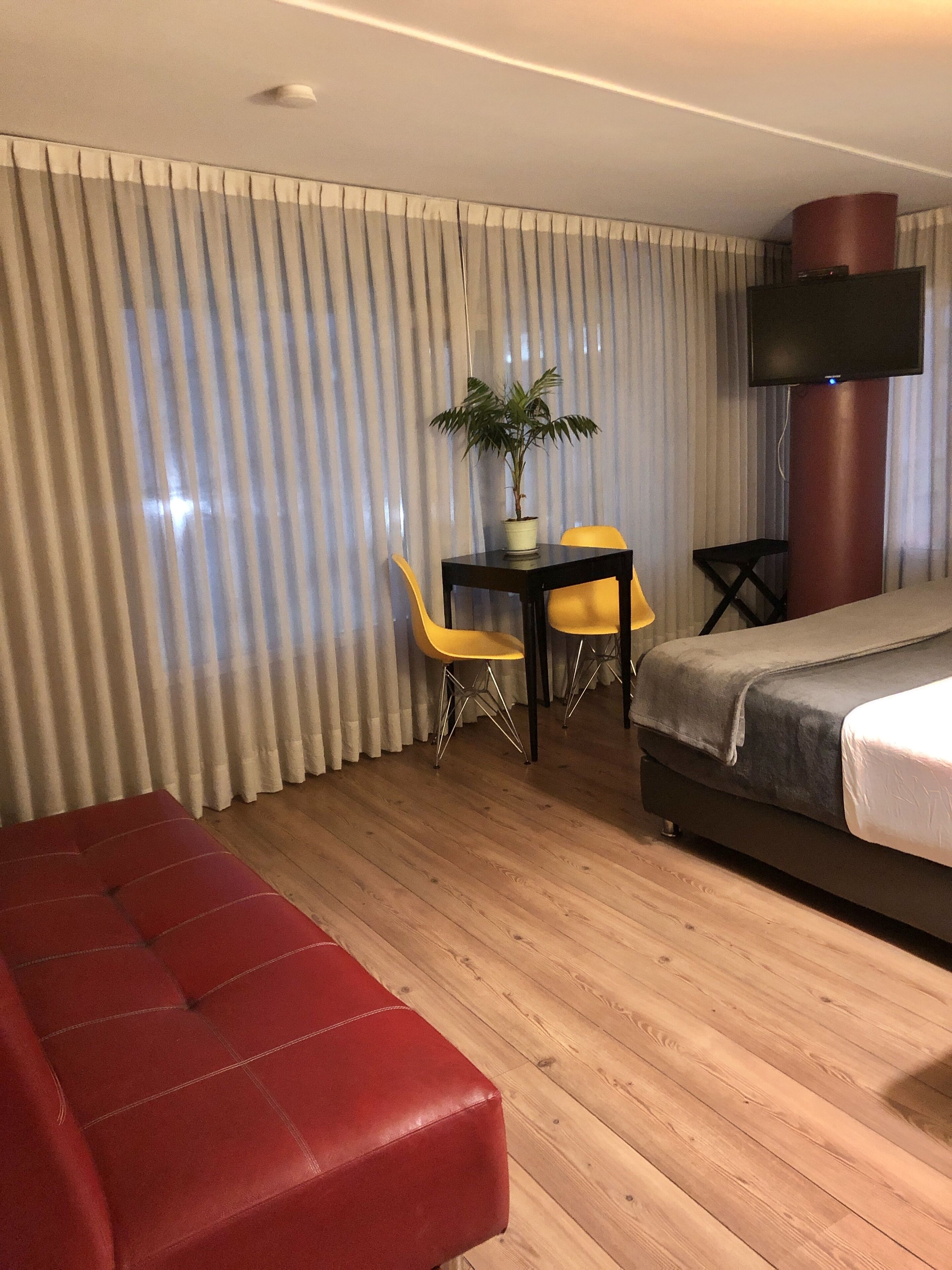 Une chambre d’hôtel avec un canapé rouge, une petite table accompagnée de deux chaises jaunes, une plante en pot, un téléviseur et un lit.