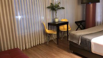Une chambre d’hôtel avec un canapé rouge, une petite table accompagnée de deux chaises jaunes, une plante en pot, un téléviseur et un lit.