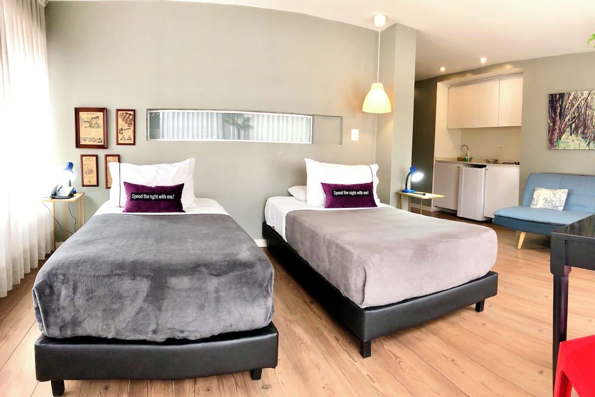 A modern hotel room with two beds, a desk, a chair, a plant, and a kitchen area.