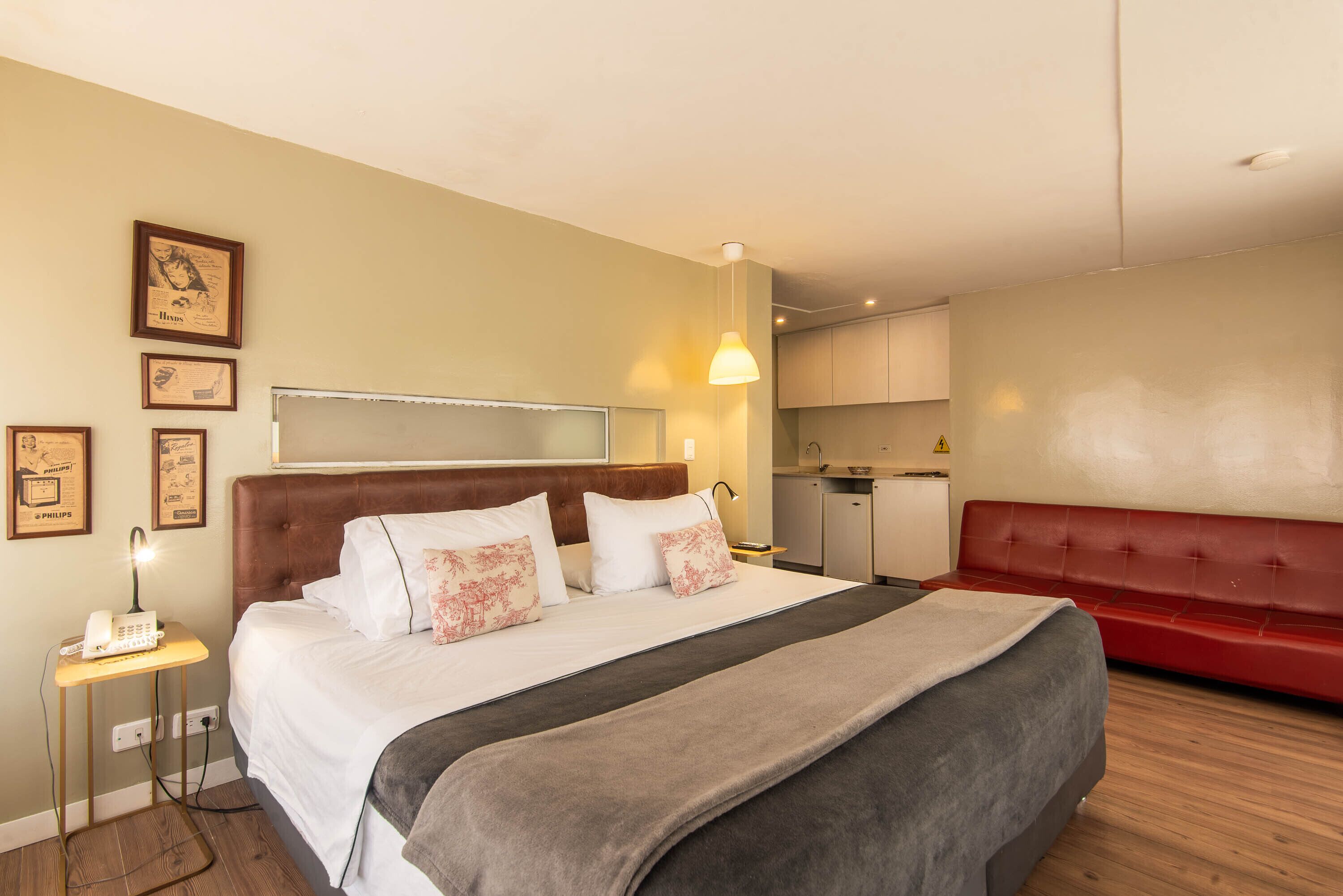 A hotel room with a bed, a red couch, a bedside table with a lamp, and a small kitchenette.