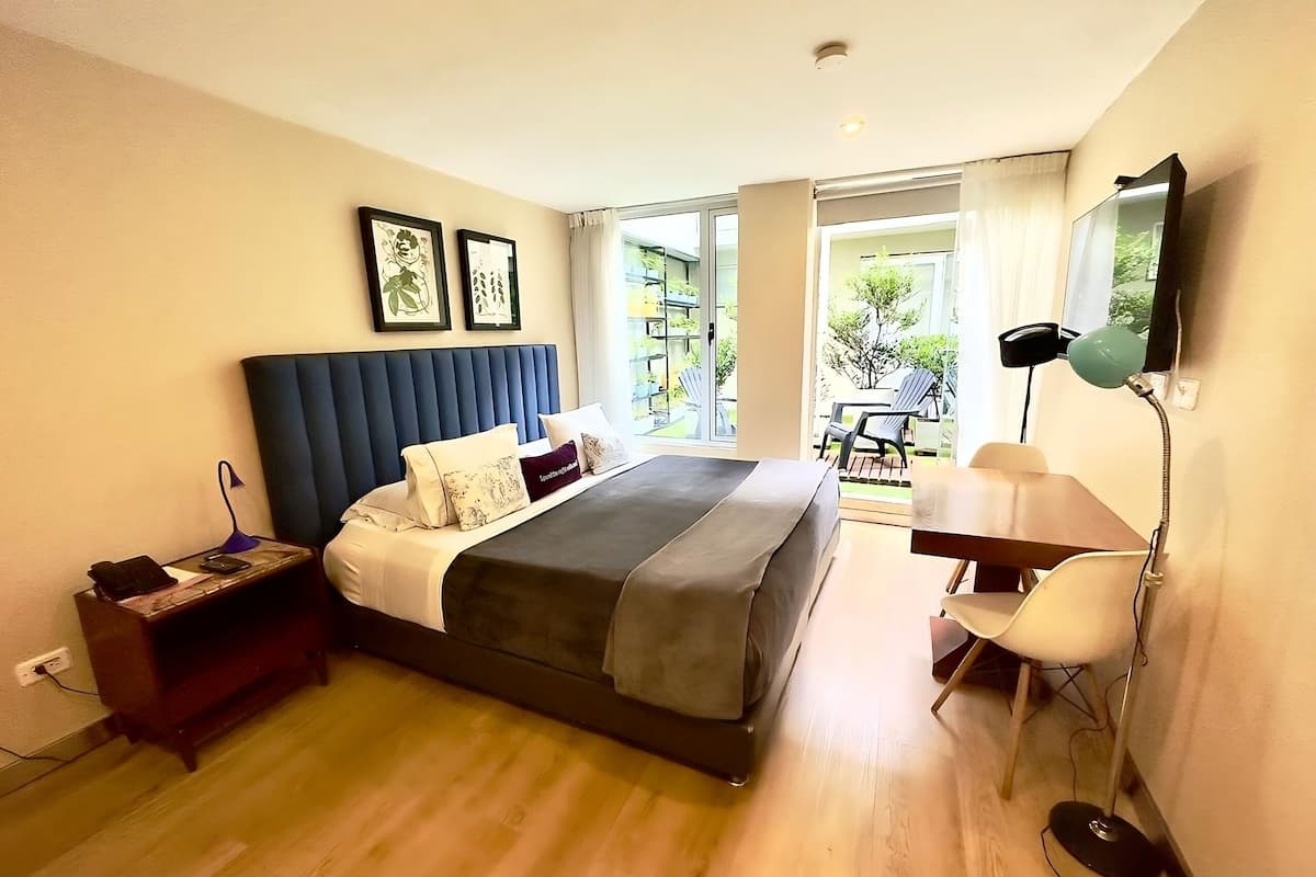 A modern bedroom with a large bed, a desk, and a chair. There is a window with a view of greenery outside.