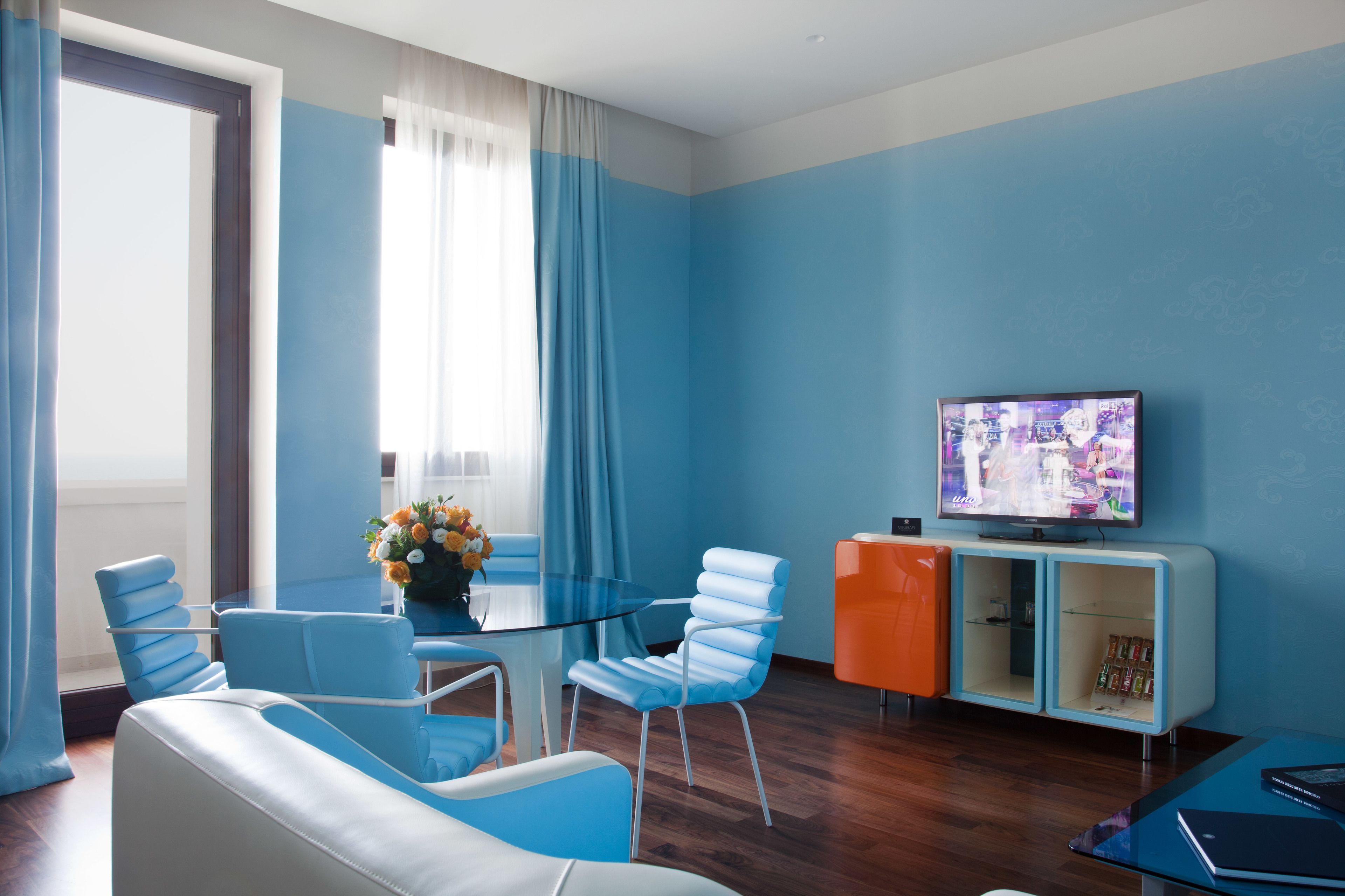 Een moderne woonkamer met een salontafel met glazen blad, blauwe en witte stoelen, een bank, een televisie op een standaard en een boekenkast.