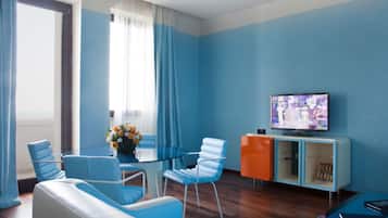 Una sala moderna con una mesa de centro de vidrio, sillas azules y blancas, un sofá, un televisor sobre un mueble bajo y un aparador con libros.