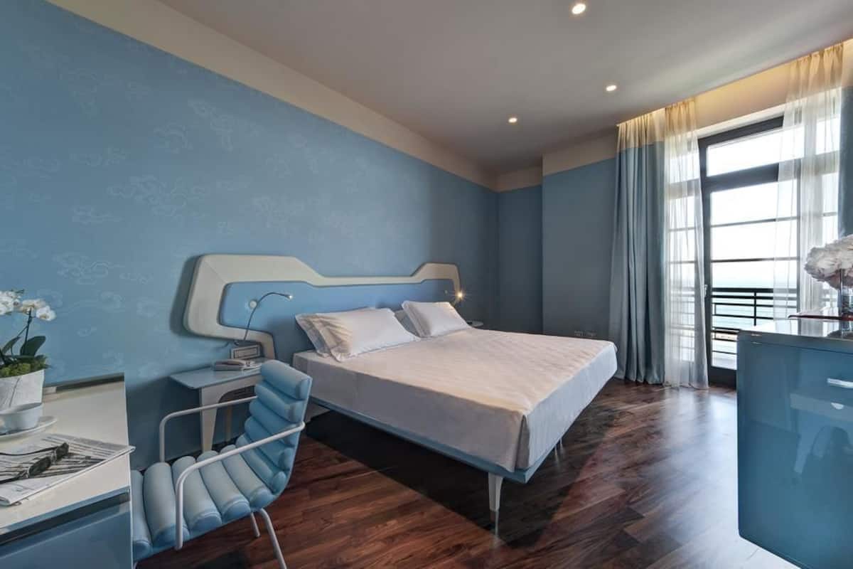 A modern hotel room with a large bed, a desk with a chair, a bedside table, and a window with curtains.