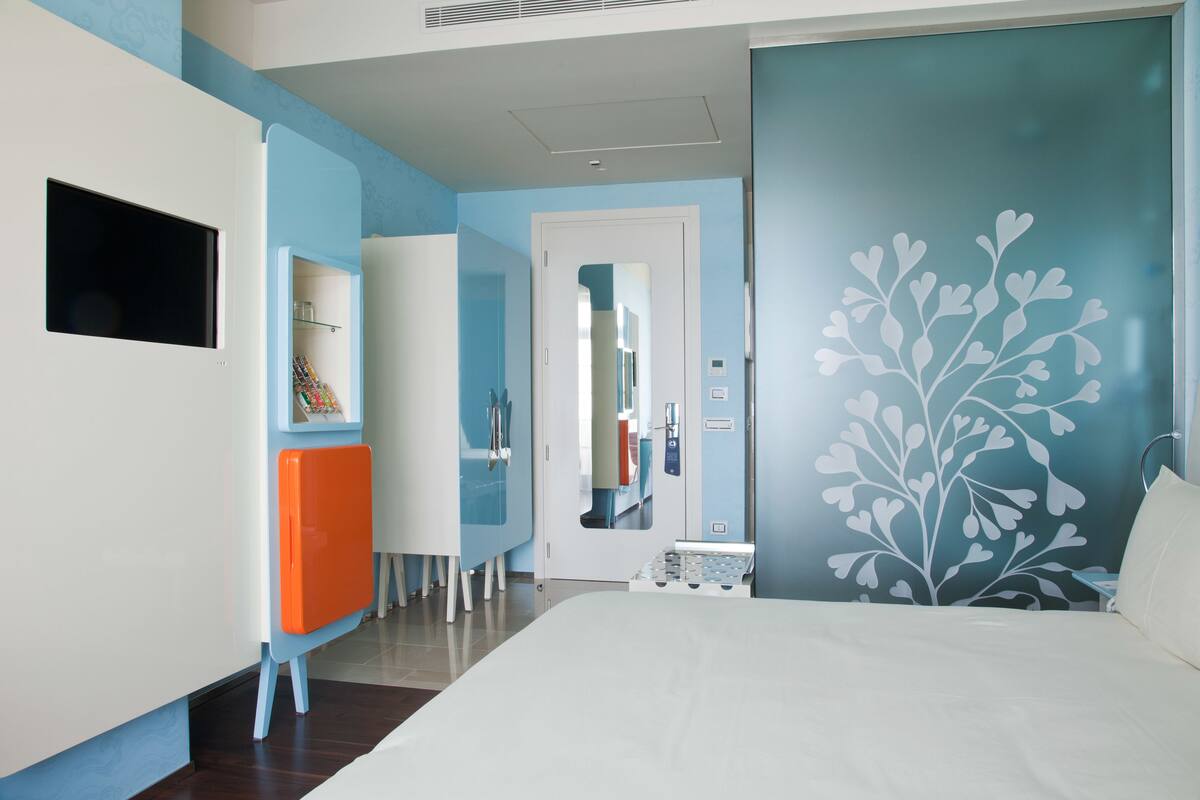 A modern hotel room with a bed, a TV, a desk, and a wardrobe.