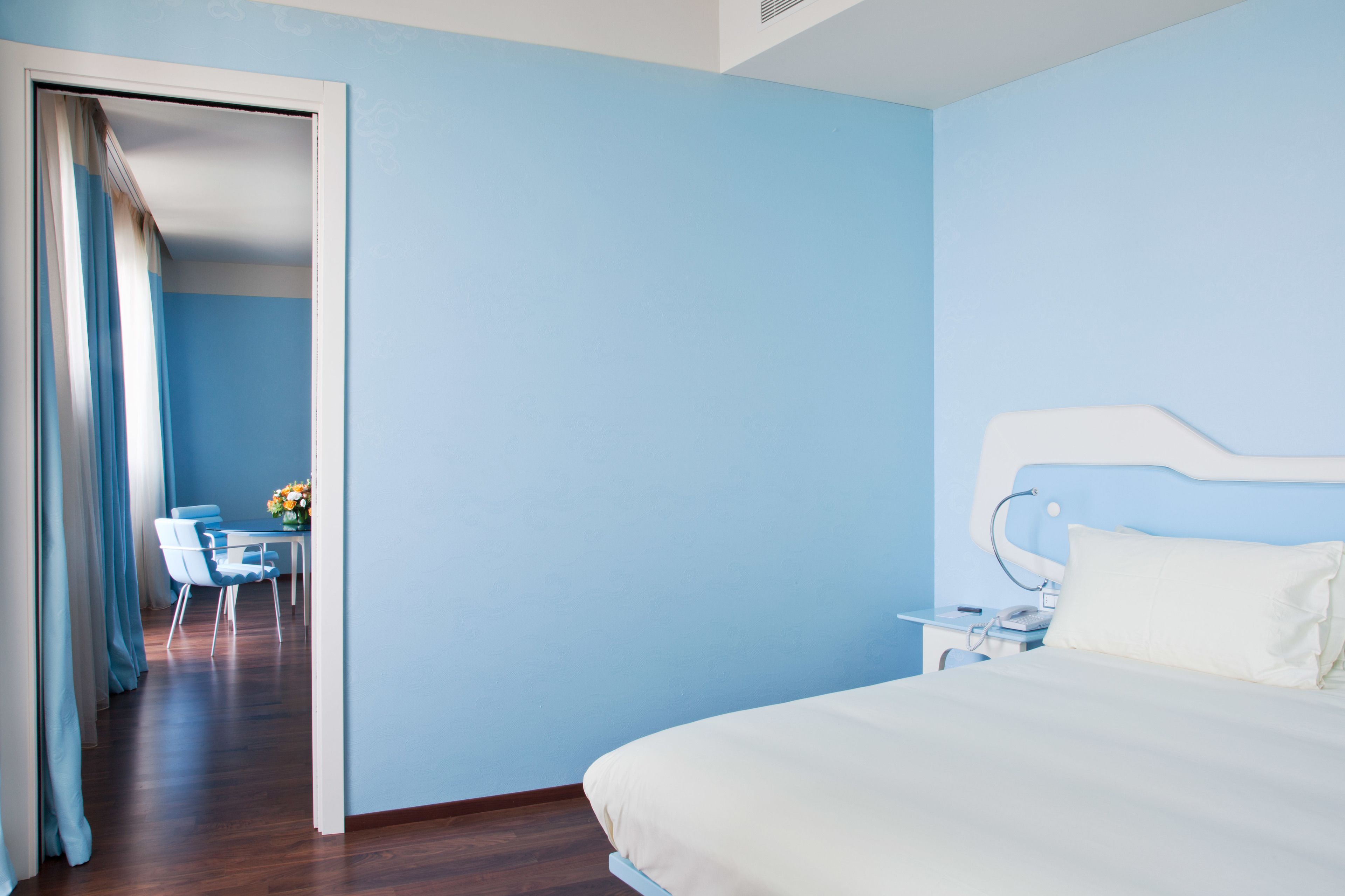 A hotel room with a bed, bedside table, and a view of a dining area with chairs and a table.