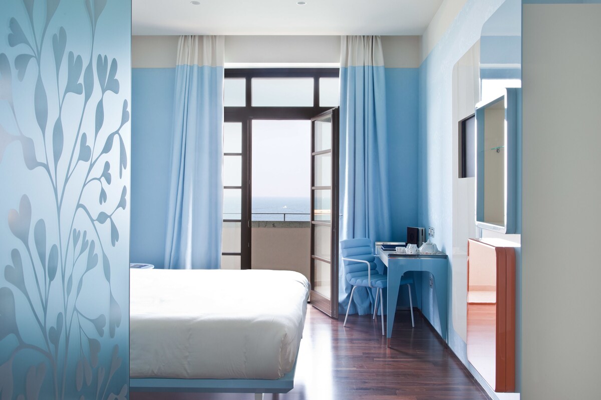Une chambre d’hôtel avec un lit, un bureau, une chaise et une vue sur la mer par la fenêtre.