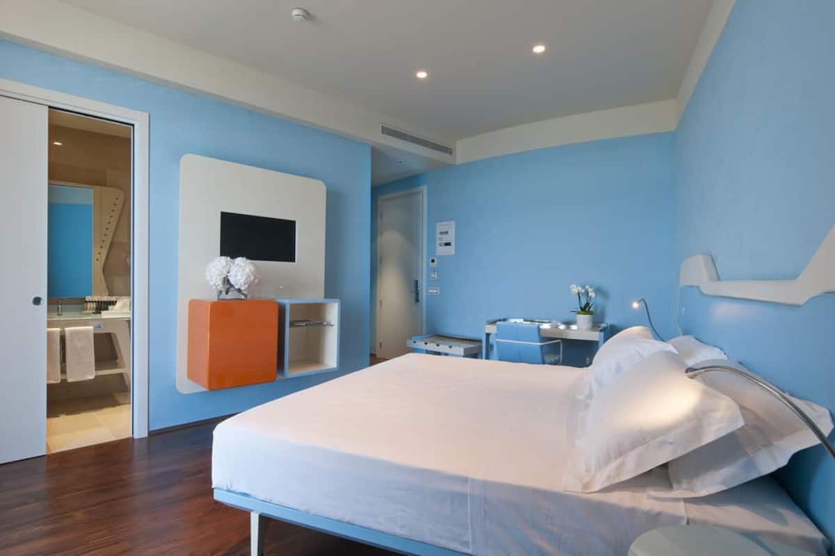 A modern hotel room with a large bed, a TV mounted on the wall, a small desk, and a bathroom visible through an open door.