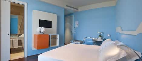 A hotel room with a bed, a TV mounted on the wall, a small desk with a chair, and a bathroom visible through an open door.