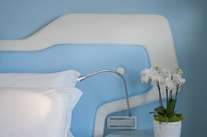 Una cama con ropa de cama blanca, un cabezal de ducha y un pequeño arreglo floral blanco en un florero.
