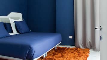 Un dormitorio con cama, cabecera blanca, ropa de cama azul, un manto naranja suave y una cortina gris.
