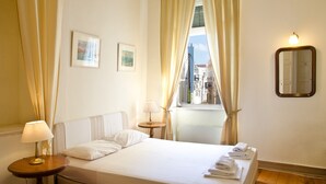 Una camera d'albergo con un letto, due lampade sui comodini, una finestra con tende, uno specchio a parete e un piccolo tavolino con lampada.