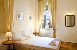 Una habitación de hotel con una cama, dos lámparas de noche, un ventanal con cortinas, un espejo colgado en la pared y una mesita con lámpara.