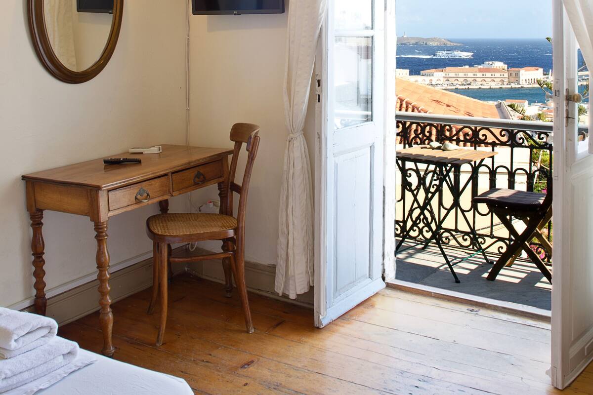 A room with a wooden desk, chair, and a bed. There is a TV mounted on the wall and a window with curtains.