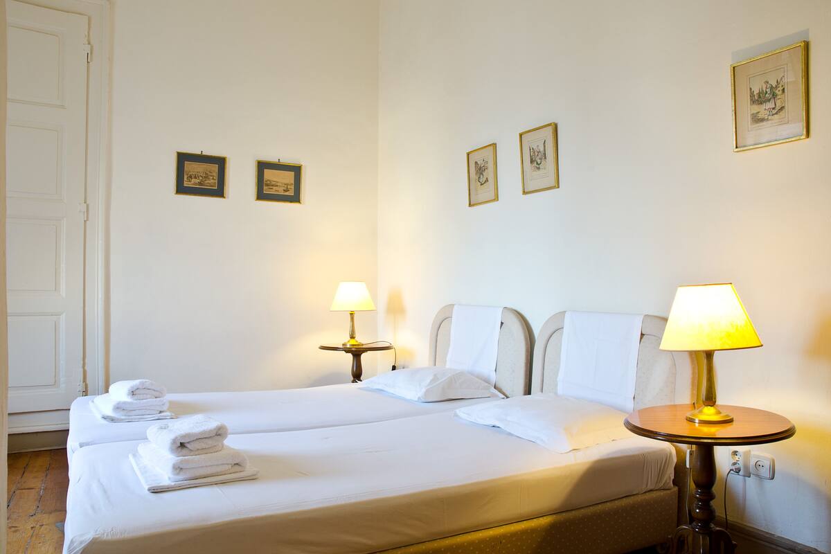 A hotel room with two beds, white linens, a round table with a lamp, and framed pictures on the wall.