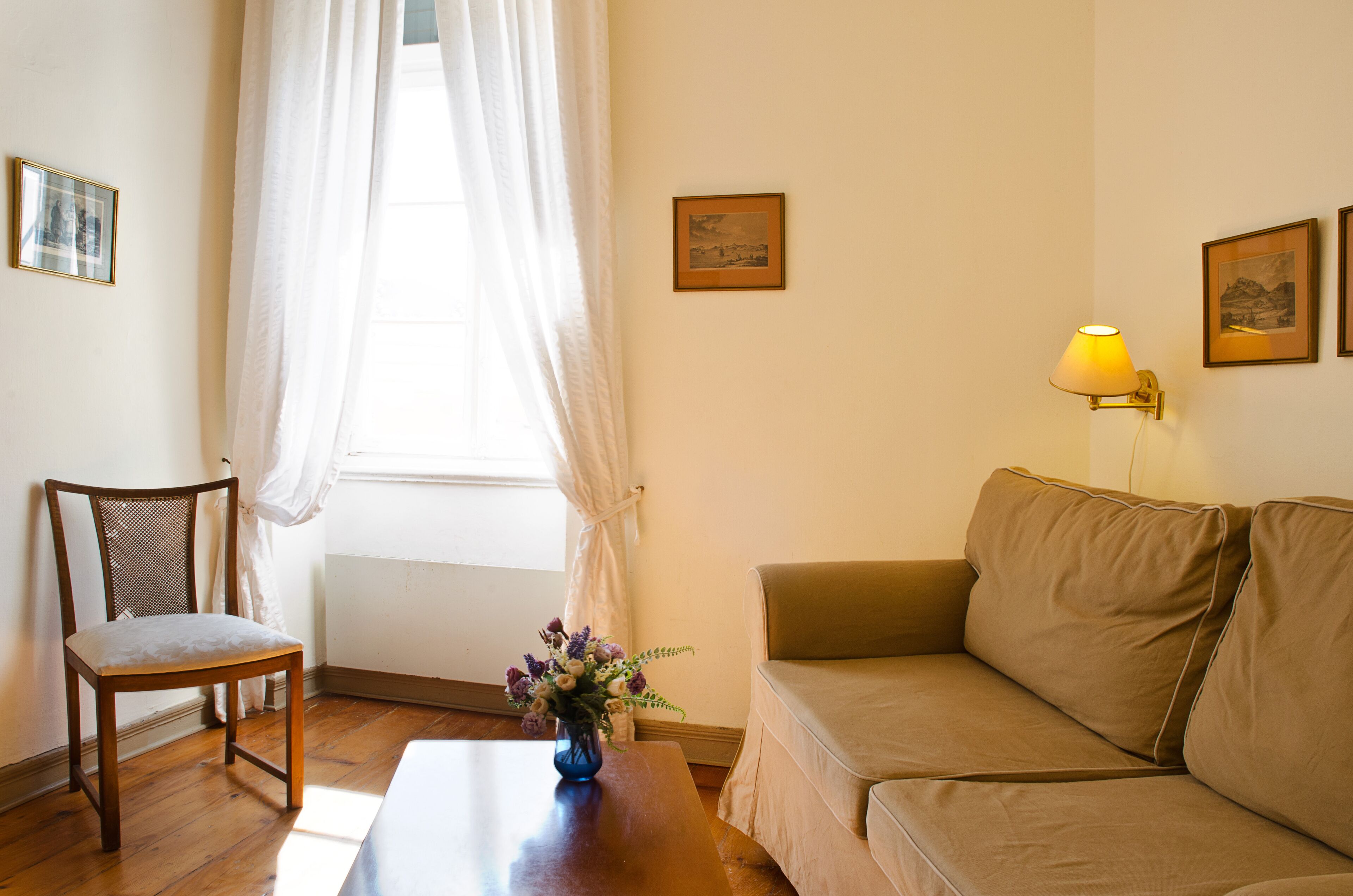 A room with a wooden chair, a beige sofa, a small table with a vase of flowers, and a window with sheer curtains.