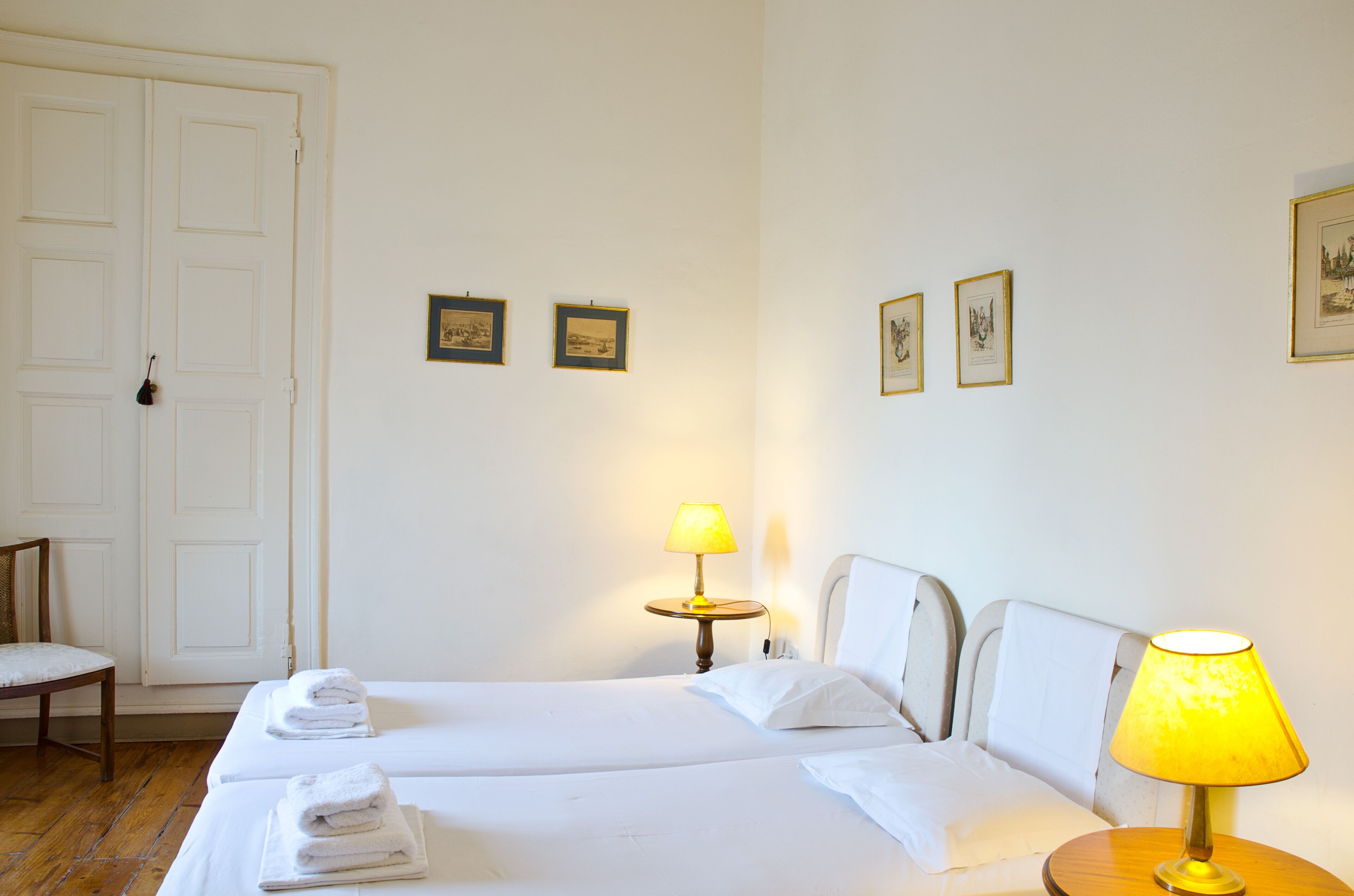 Una habitación de hotel con dos camas, una silla, una lámpara y cuadros en la pared.